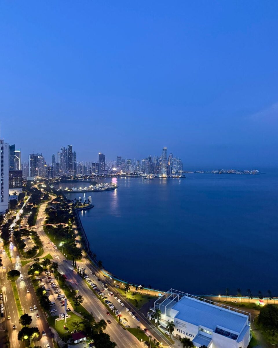 Nighttime aerial view of a coastal city skyline with illuminated streets and high-rise buildings reflecting on calm water.