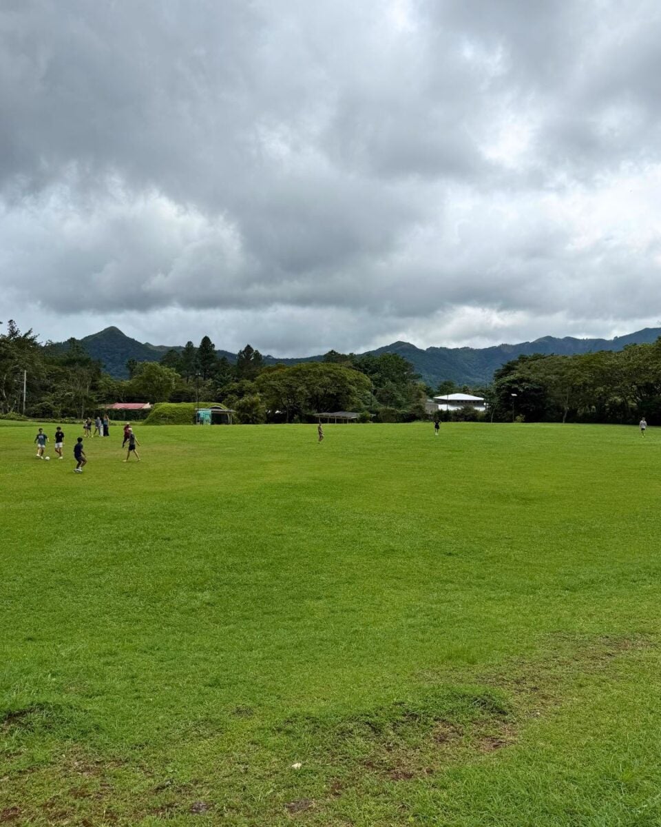 Children playing on a large grassy field under a cloudy sky with mountains in the background.