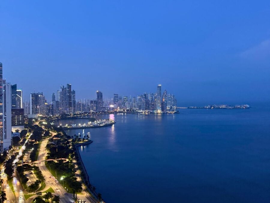 Nighttime aerial view of a coastal city skyline with illuminated streets and high-rise buildings reflecting on calm water.