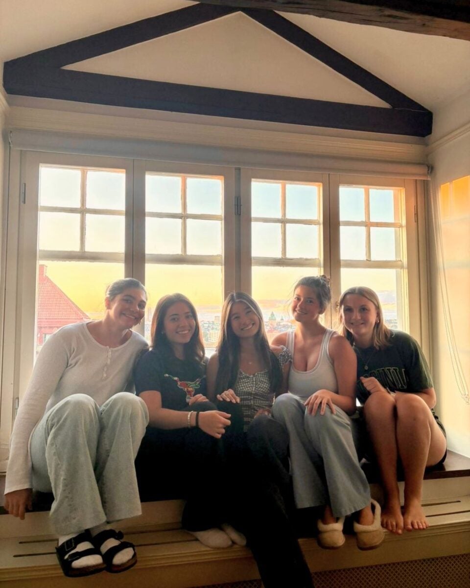 Five young women sitting closely together on a wide windowsill with sunlight streaming through the multi-pane window behind them.