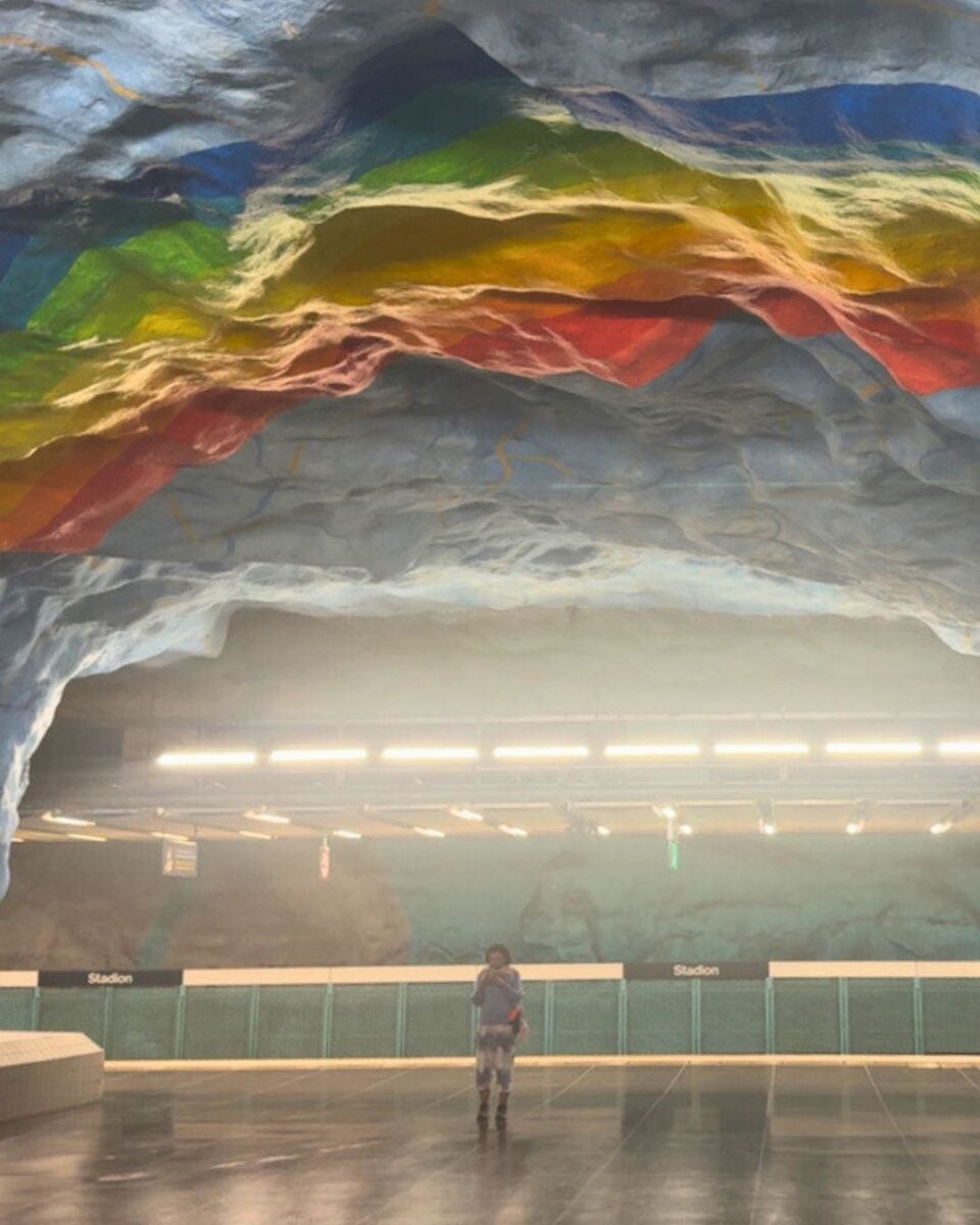 Colorful rainbow patterns painted on the rocky ceiling of an underground subway station with a lone person standing on the platform.