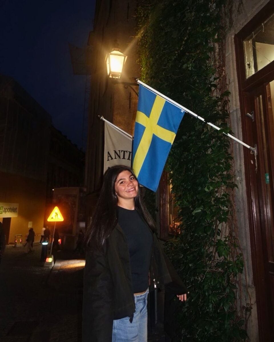 Smiling woman standing under a Swedish flag mounted on a building at night on a dimly lit street.