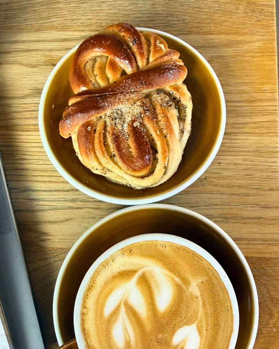 Freshly baked cardamom bun with sugar topping served in a bowl alongside a latte with leaf-shaped foam art on a wooden table.