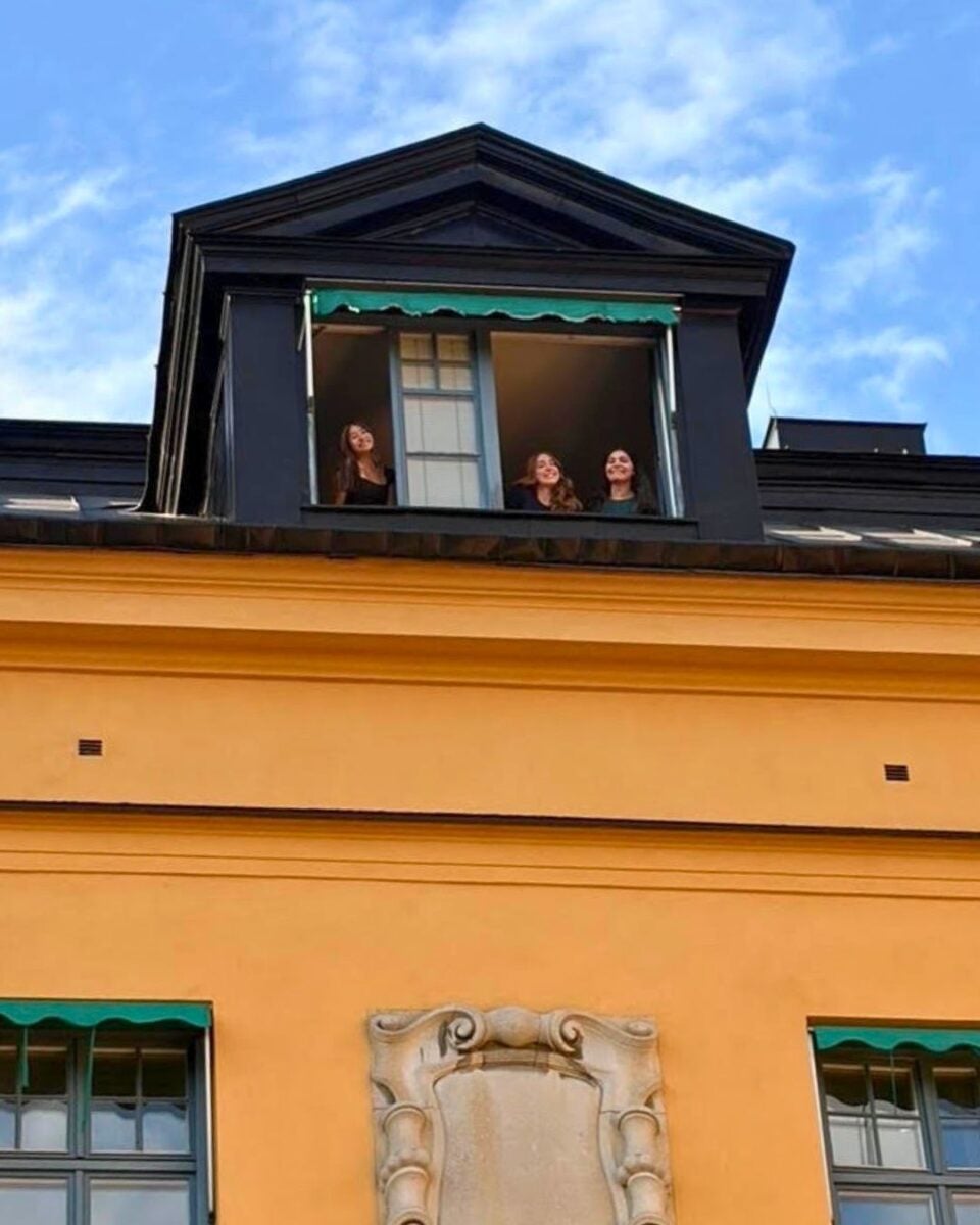 Three people smiling and looking out from a black dormer window on the top floor of a yellow building under a blue sky.