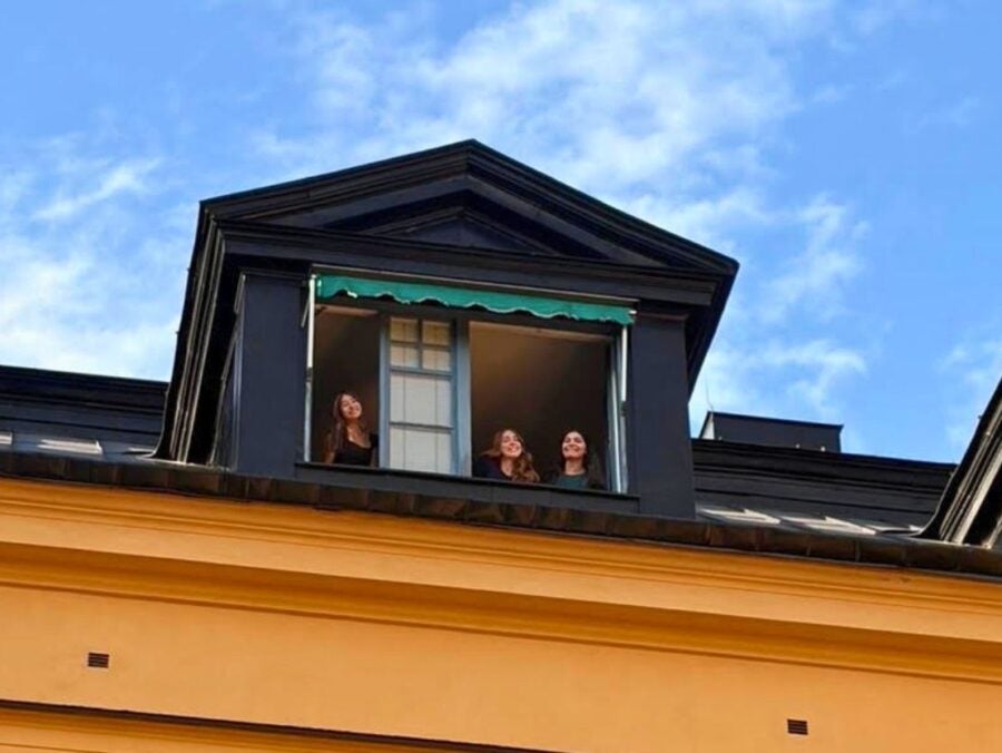 Three people smiling and looking out from a black dormer window on the top floor of a yellow building under a blue sky.